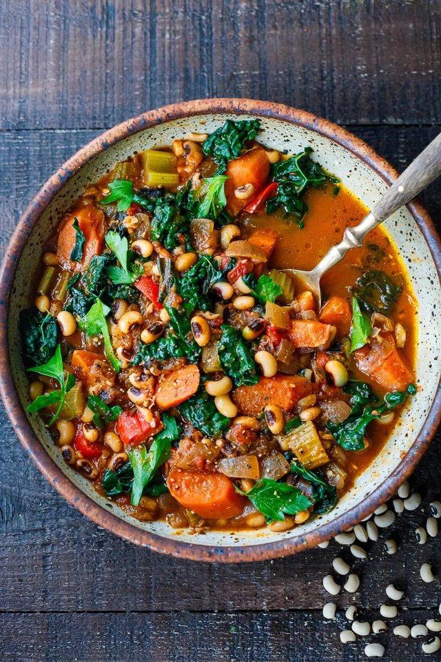 A speckled bowl of black-eyed pea and kale stew with carrots, celery, red pepper and parsley, with a spoon resting in it